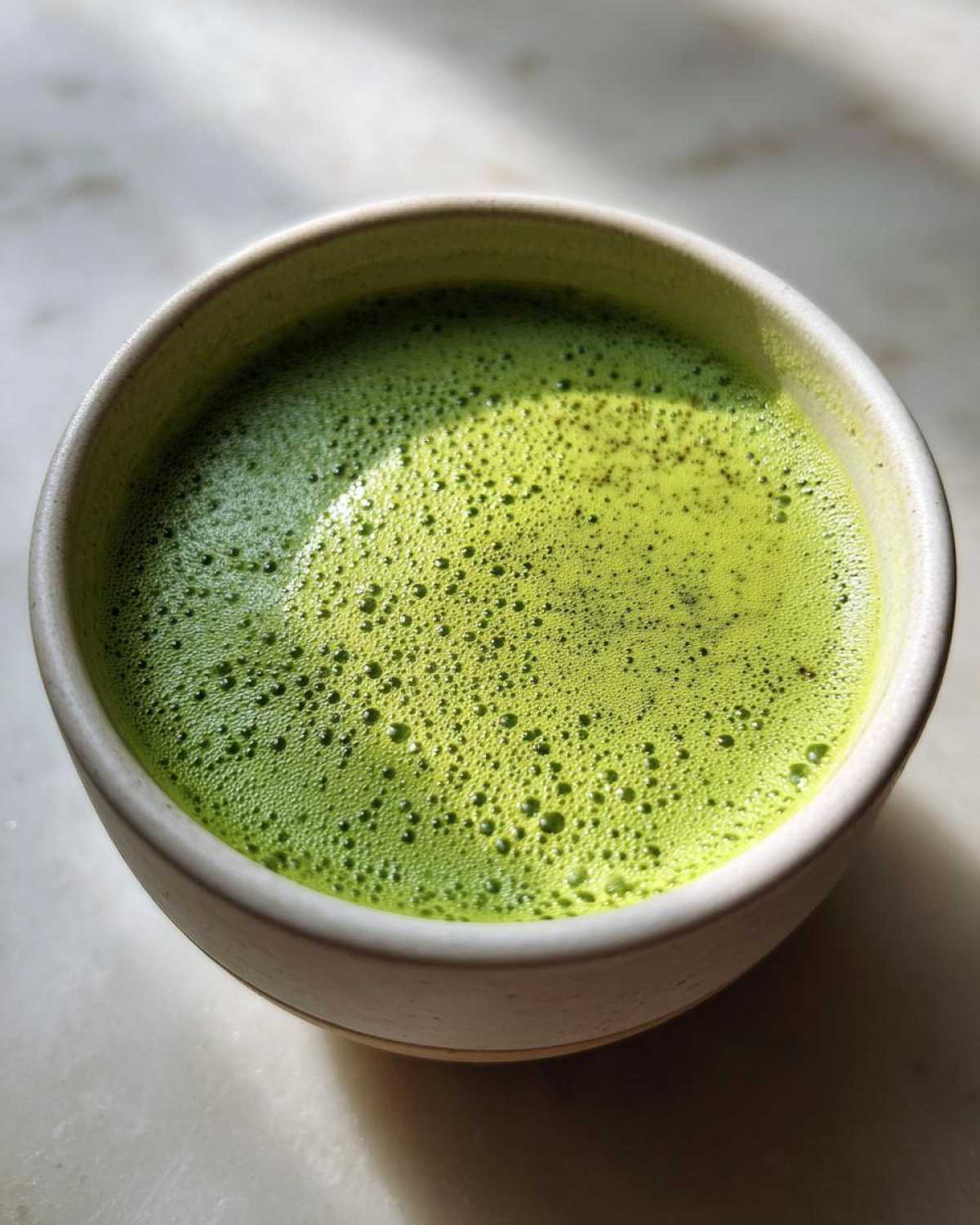 Close-up of a vibrant green, foamy beverage, likely matcha, served in a light-colored ceramic bowl, unrelated to asparagus soup.