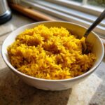 A close-up of vibrant yellow turmeric rice served in a white bowl with a spoon resting inside.