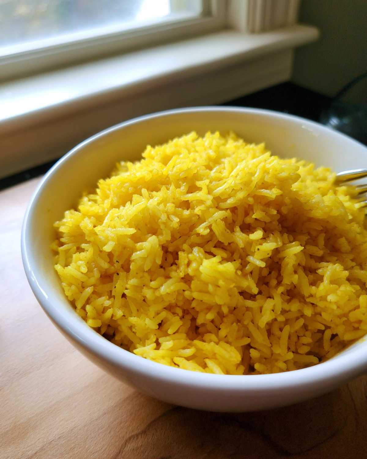 Close-up of a white bowl filled with fluffy, bright yellow turmeric rice, set near a window.