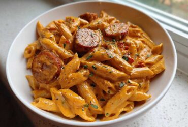A close-up of creamy Cajun sausage pasta featuring penne coated in an orange sauce with sliced sausage and parsley.