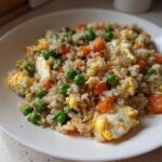 Close-up of a white plate filled with savory cauliflower rice mixed with scrambled egg, bright green peas, and diced carrots.