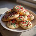 A pile of soft, round christmas sprinkle sugar cookies topped with red, white, and green sprinkles on a white plate.