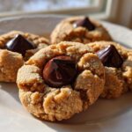 Four classic peanut butter blossom cookies with a melted chocolate kiss pressed into the center, sitting on a white plate.