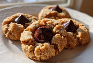 Four classic peanut butter blossom cookies with a melted chocolate kiss pressed into the center, sitting on a white plate.