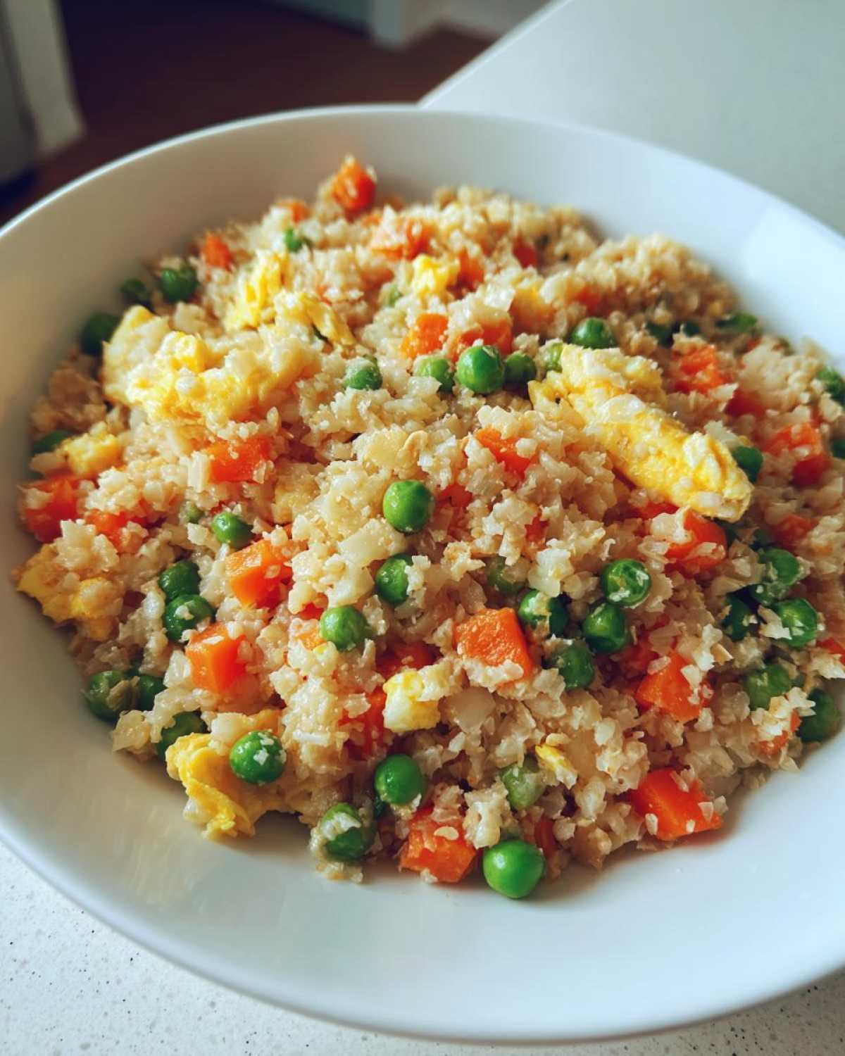 Close-up of a bowl of vibrant cauliflower rice mixed with bright green peas and diced carrots, and scrambled egg pieces.