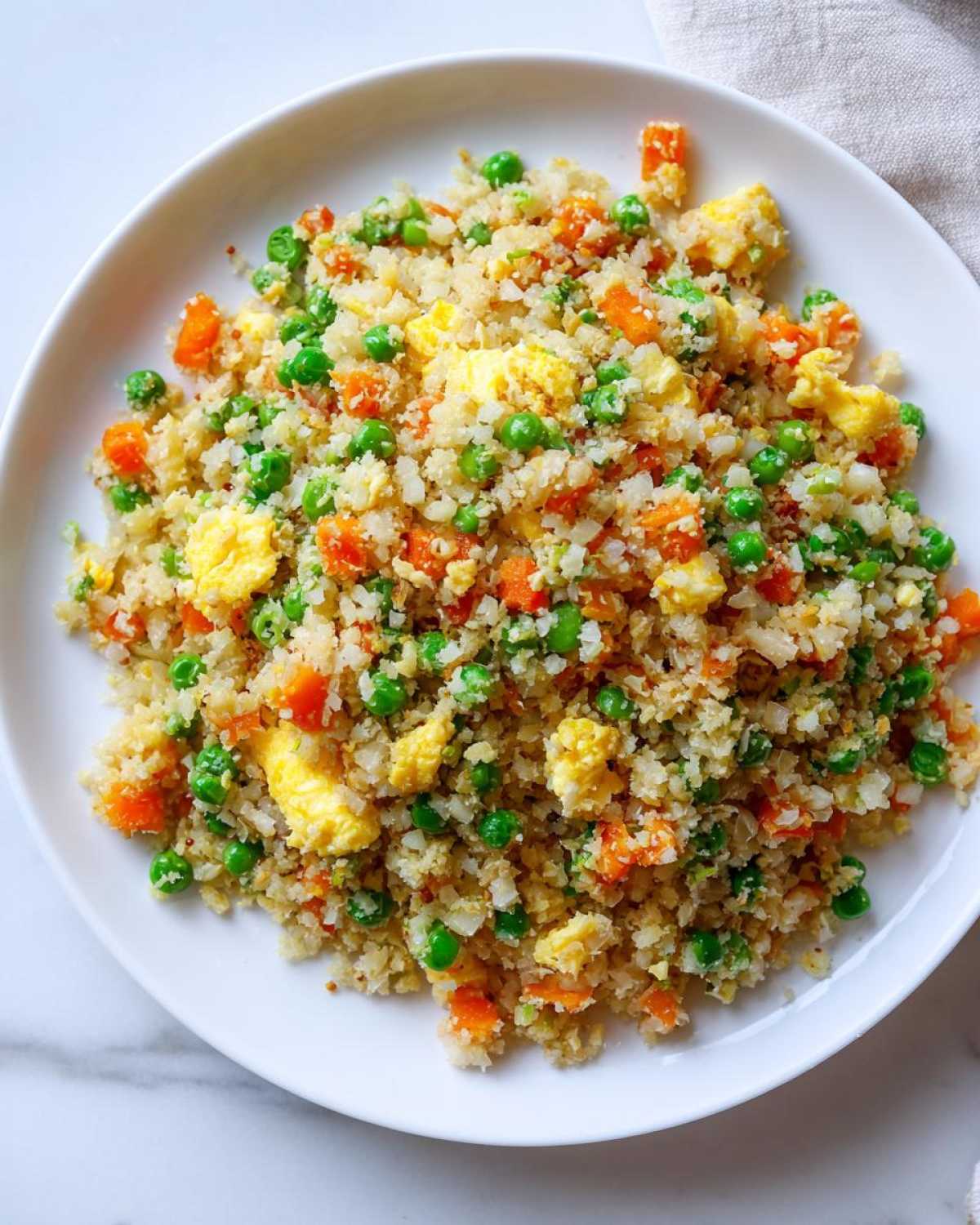 Overhead view of a white plate filled with bright cauliflower rice mixed with green peas and scrambled egg pieces.