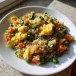 A white plate filled with vibrant cauliflower rice mixed with scrambled egg pieces, green peas, and diced carrots.
