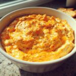 A close-up of creamy, orange beer cheese dip topped with black pepper in a white bowl, with crackers visible in the background.