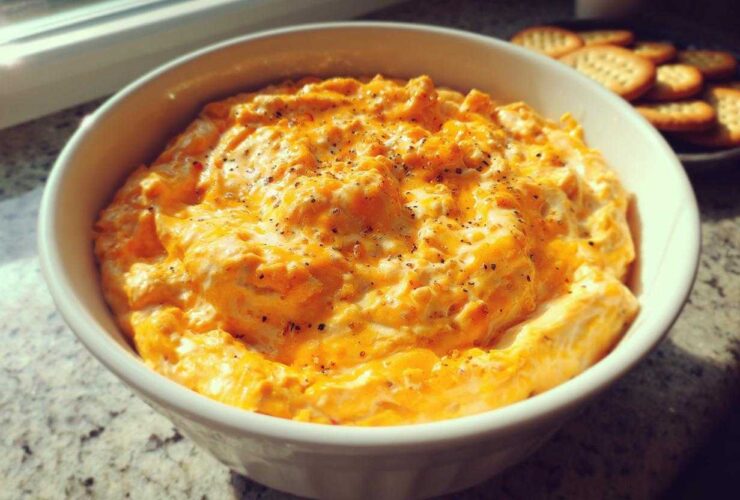A close-up of creamy, orange beer cheese dip topped with black pepper in a white bowl, with crackers visible in the background.
