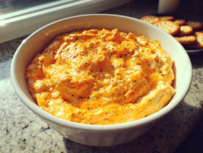 A close-up of creamy, orange beer cheese dip topped with black pepper in a white bowl, with crackers visible in the background.