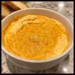 Close-up of a thick, orange-hued creamy beer cheese dip served in a white bowl, with toasted bread slices visible in the background.