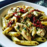 Close-up of creamy chicken pesto pasta featuring penne, sliced chicken breast, and sun-dried tomatoes.