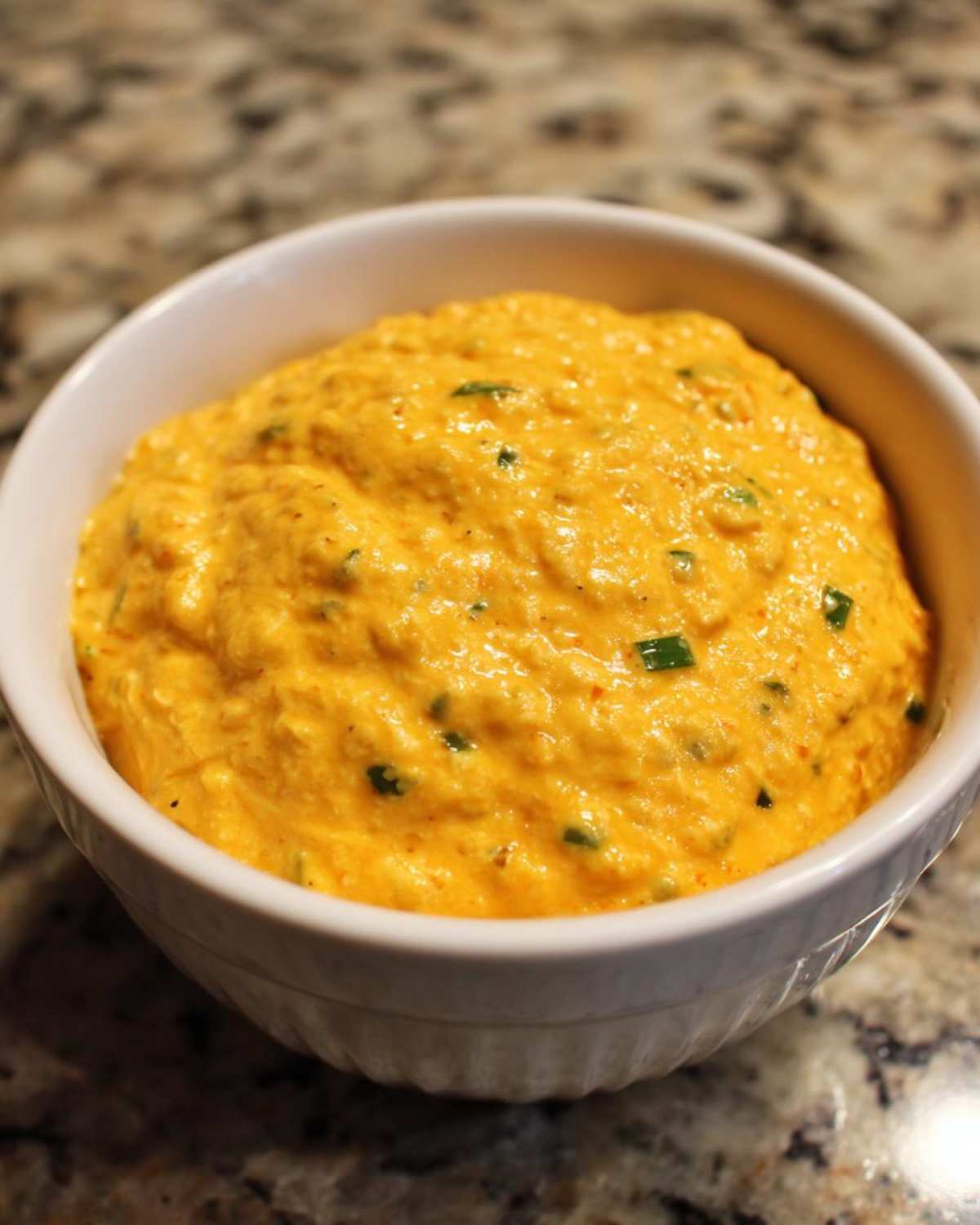 A close-up of thick, bright orange cottage cheese queso dip speckled with green chives in a white bowl.