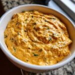 Close-up of thick, creamy orange cottage cheese queso mixed with green herbs, served in a white bowl.