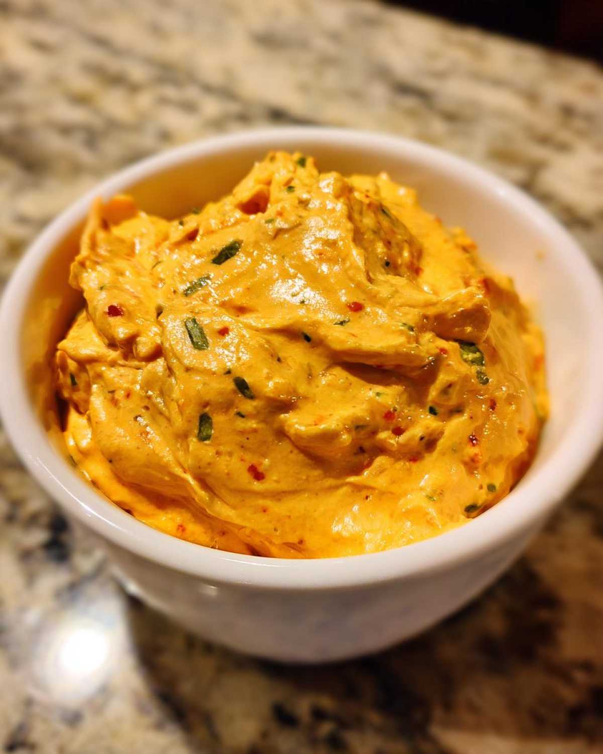 A close-up of thick, orange-yellow cottage cheese queso dip speckled with green herbs and red pepper flakes in a white bowl.