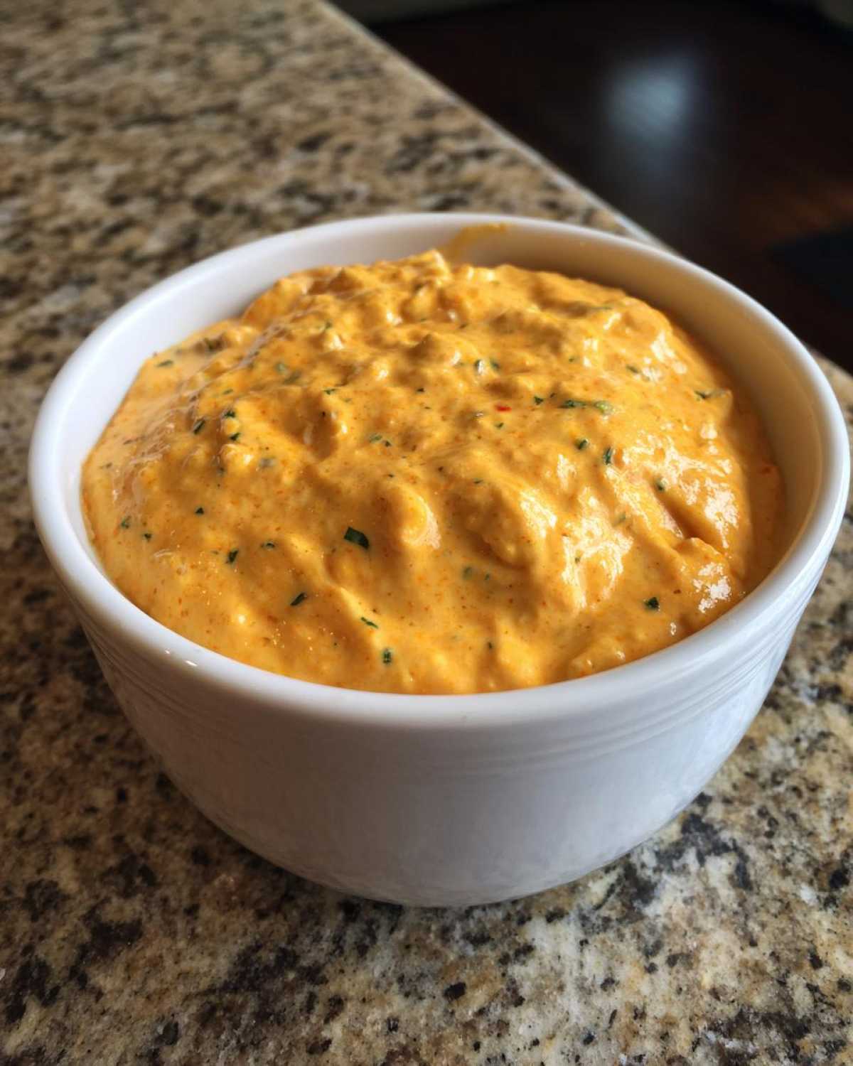 A close-up of thick, orange cottage cheese queso dip speckled with green herbs in a white bowl.