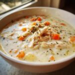 Close-up of a white bowl filled with homemade cream of chicken soup, featuring shredded chicken, carrots, and herbs.