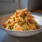 A generous serving of creamy, orange-hued kani salad mixed with thin strips of green cucumber in a white bowl.