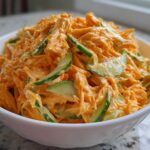 A close-up view of a white bowl filled with vibrant orange kani salad mixed with creamy dressing and slices of green cucumber.
