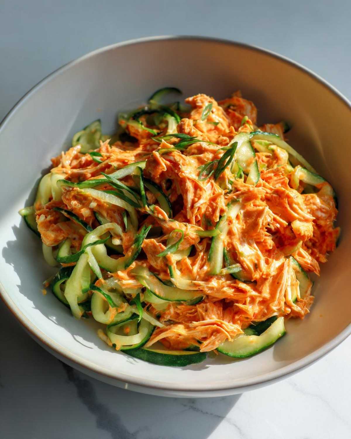 A bowl filled with creamy, orange-tinted kani salad mixed with thin ribbons of fresh cucumber and topped with green onions.