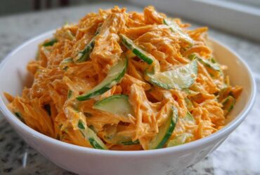 A close-up view of a white bowl filled with vibrant orange kani salad mixed with creamy dressing and slices of green cucumber.
