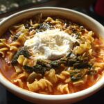 A close-up of a bowl of creamy lasagna soup featuring pasta, spinach, and topped with a dollop of ricotta and shredded Parmesan cheese.