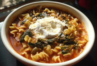 A close-up of a bowl of creamy lasagna soup featuring pasta, spinach, and topped with a dollop of ricotta and shredded Parmesan cheese.