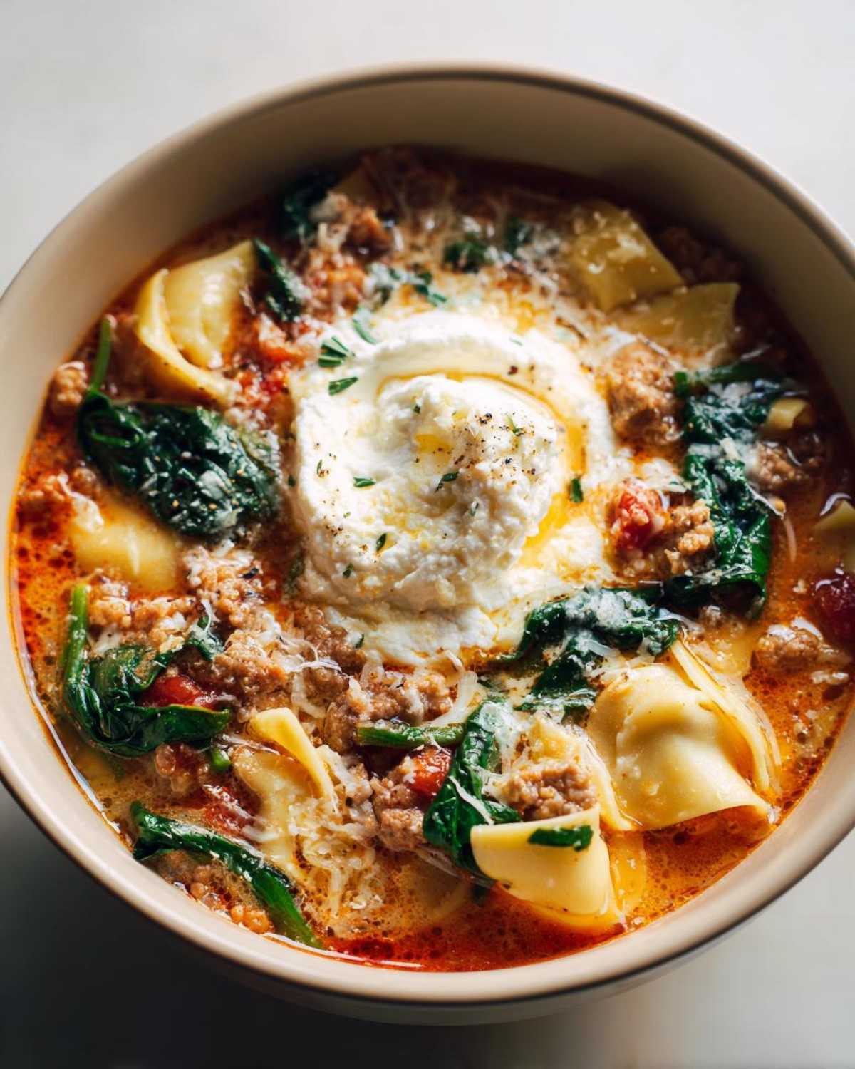 Close-up of a hearty bowl of creamy lasagna soup with sausage, spinach, pasta, and a dollop of ricotta cheese.
