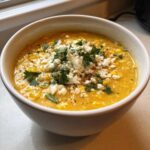 A close-up of a white bowl filled with creamy yellow Mexican street corn soup, topped with crumbled white cheese and fresh cilantro.