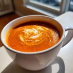 A close-up of vibrant orange roasted tomato soup topped with a swirl of cream in a white mug.