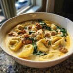 A close-up of a white bowl filled with creamy sausage tortellini soup, featuring chunks of sausage and wilted spinach.