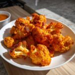 A close-up of crispy, bright orange buffalo cauliflower florets piled on a white plate, with a side of dipping sauce visible.