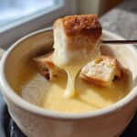 A piece of toasted bread is dipped into a creamy, yellow cheese fondue using a fondue fork.