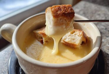 A piece of toasted bread is dipped into a creamy, yellow cheese fondue using a fondue fork.