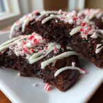 A close-up view of a double chocolate peppermint cookie broken in half, drizzled with white chocolate and topped with crushed candy canes.