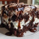 A decadent slice of hot chocolate poke cake showing dark chocolate cake layers filled with white cream and drizzled heavily with chocolate fudge sauce.