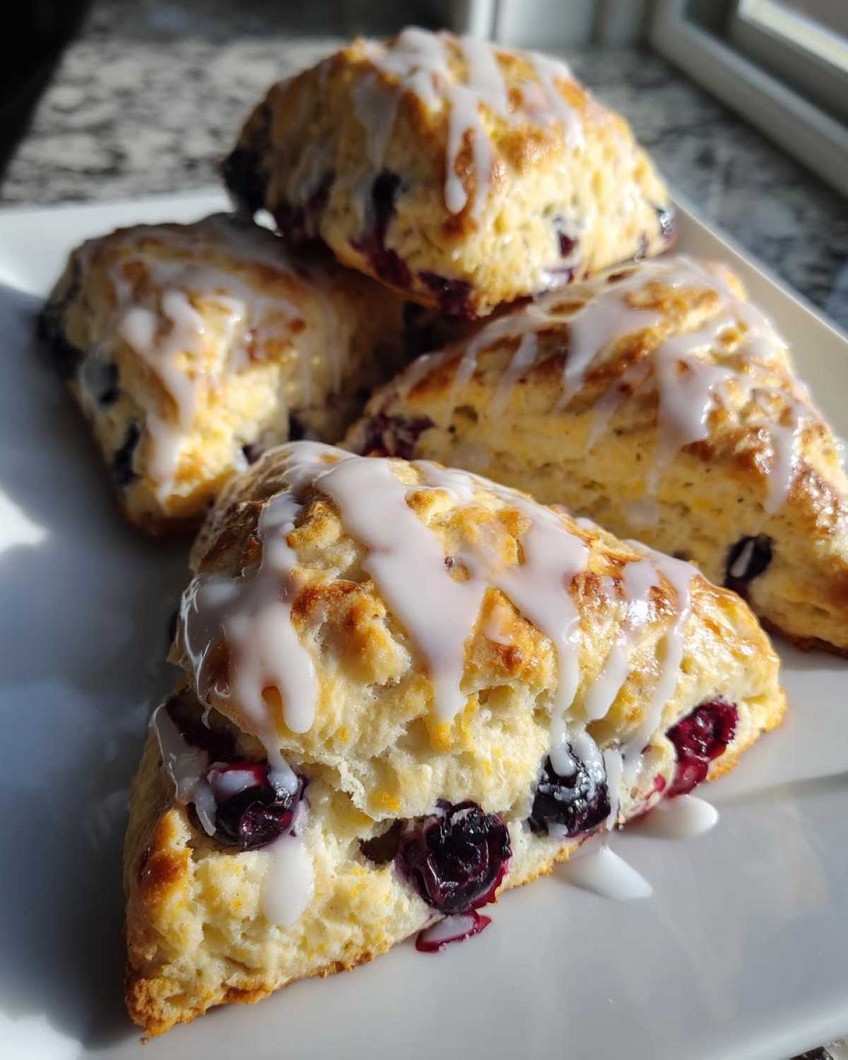Four flaky blueberry scones drizzled with white vanilla glaze stacked on a white plate.