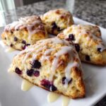 Four flaky blueberry scones drizzled generously with vanilla glaze, served on a white plate.
