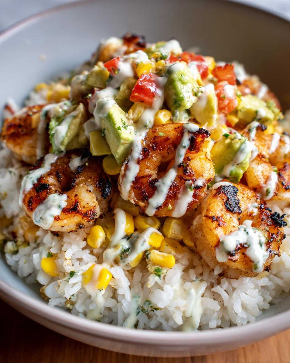 Close-up of a delicious shrimp bowl featuring grilled shrimp, white rice, corn, avocado, and a creamy white drizzle.