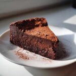 A decadent slice of flourless chocolate torte, heavily dusted with cocoa powder, sitting on a white plate.