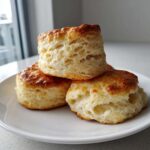 Three tall, fluffy scones stacked on a white plate, showing their flaky interior and golden-brown tops.