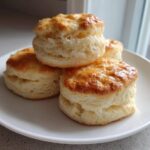 A stack of four perfectly golden and fluffy scones resting on a white plate near a window.
