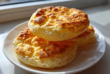Three golden-brown, fluffy cloud bread rounds stacked on a white plate near a window.