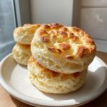 A stack of three golden-brown, airy cloud bread rounds resting on a rustic white plate near a window.