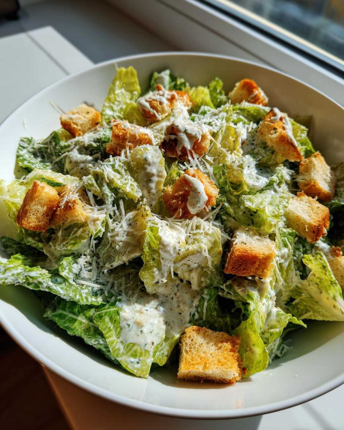 Close-up of a fresh caesar salad in a white bowl, topped with creamy dressing, grated parmesan, and golden croutons.