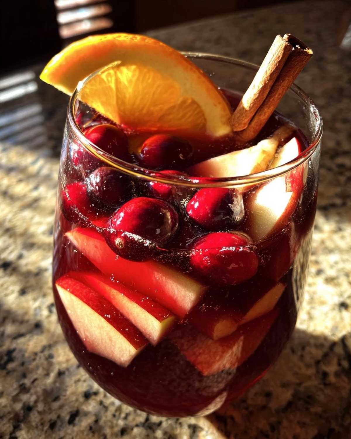 A glass filled with rich red holiday sangria, loaded with cranberries, apple slices, an orange wedge, and a cinnamon stick.