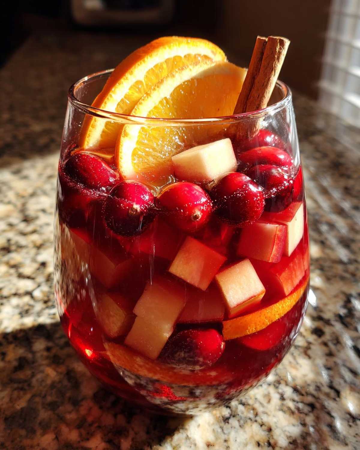 A glass filled with vibrant red holiday sangria, loaded with cranberries, apple chunks, orange slices, and a cinnamon stick.