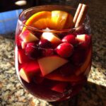 Close-up of a glass filled with rich red holiday sangria, loaded with apple chunks, cranberries, and an orange slice, garnished with a cinnamon stick.