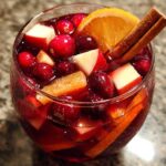 Close-up of a glass filled with rich red holiday sangria, loaded with cranberries, apple chunks, and an orange slice garnished with cinnamon sticks.