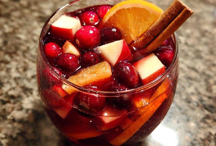 Close-up of a glass filled with rich red holiday sangria, loaded with cranberries, apple chunks, and an orange slice garnished with cinnamon sticks.